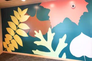 A wall mural featuring large, stylized leaves in shades of yellow, green, brown, pink, and white on a teal background. A security camera is mounted near the top right corner.