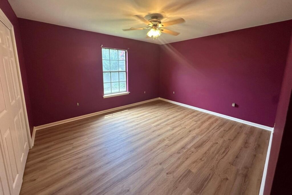 An empty room with purple walls, light wood flooring, a ceiling fan with lights, and a single window letting in natural light. A white closet door is visible on the left.