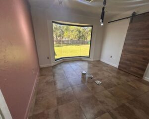 A mostly empty room with tiled floors, a large bay window overlooking a green backyard, a pink accent wall, a wooden sliding barn door, and a few paint cans and supplies on the floor.