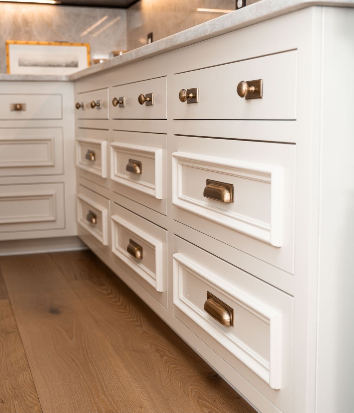 White kitchen cabinets with brass handles and knobs, set on a light wood floor. The counter above is light-colored, and a framed picture is partially visible in the background. The setting appears modern and clean.