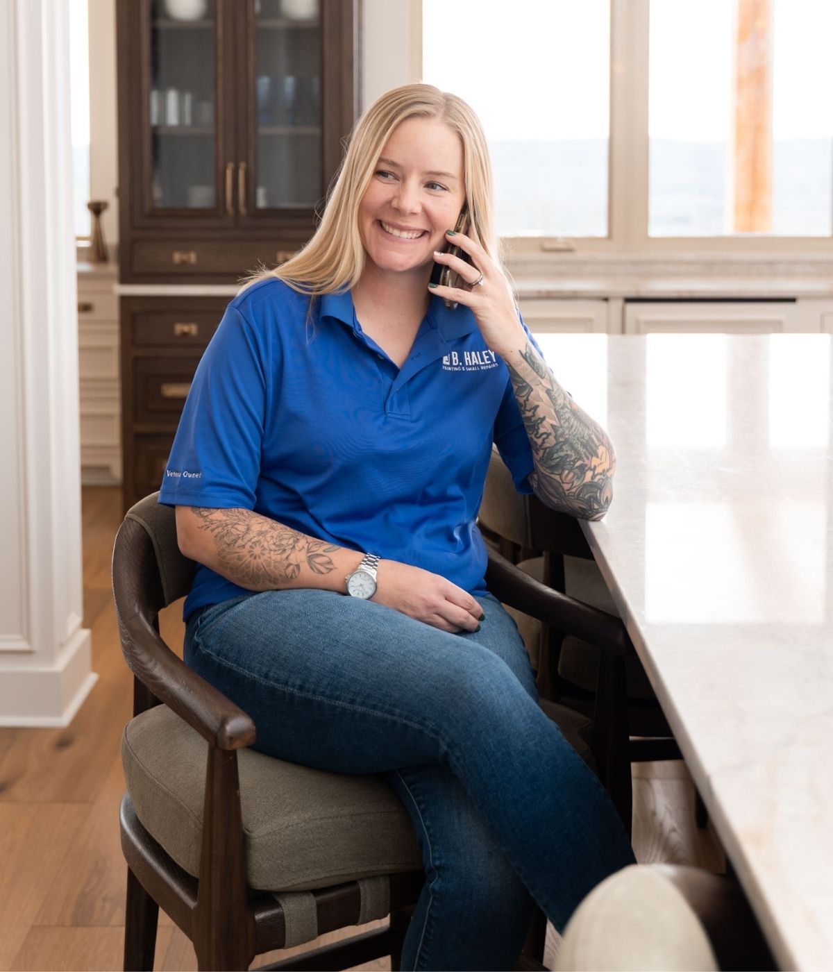 A smiling person with long blonde hair, tattoos on both arms, and a blue polo shirt sits on a chair at a kitchen counter, talking on a cellphone. Bright daylight streams through large windows behind them.