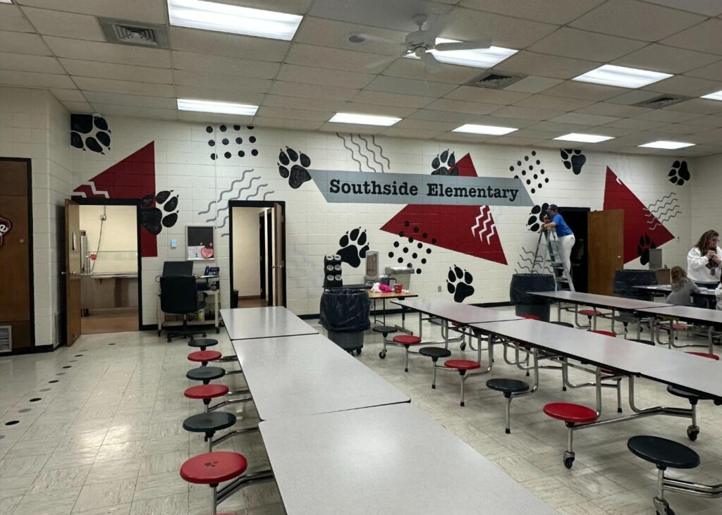 An elementary school cafeteria with long tables and red stools. The wall features Southside Elementary in bold letters with red arrows, black paw prints, and zigzag patterns. Two people are working near a ladder.