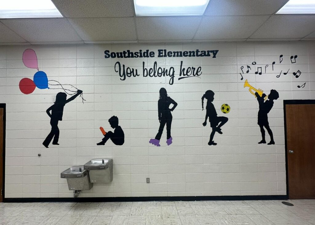 A mural on a school wall shows five silhouettes of children: one with balloons, one reading, one standing with hands on hips, one kicking a soccer ball, and one playing a trumpet. Text reads Southside Elementary You belong here.