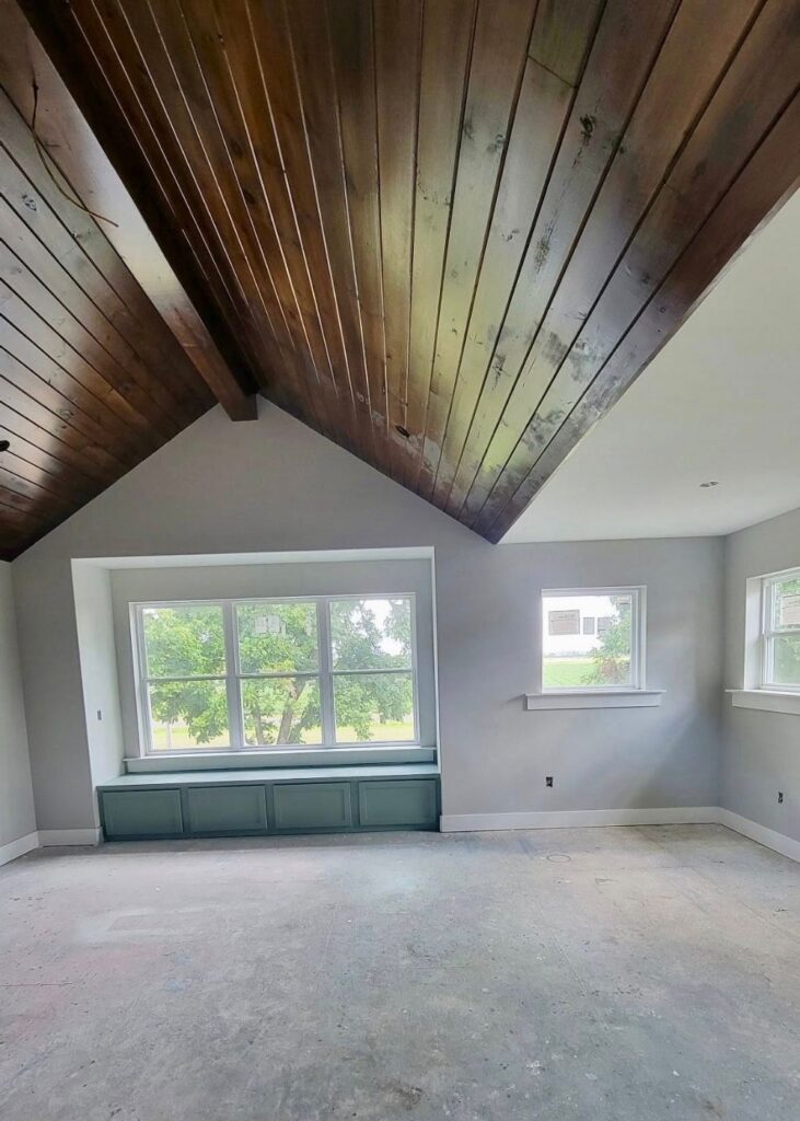A bright, empty room with a wooden vaulted ceiling, large windows with green window seats, light gray walls, and unfinished floors. Trees and a house are visible outside the windows.