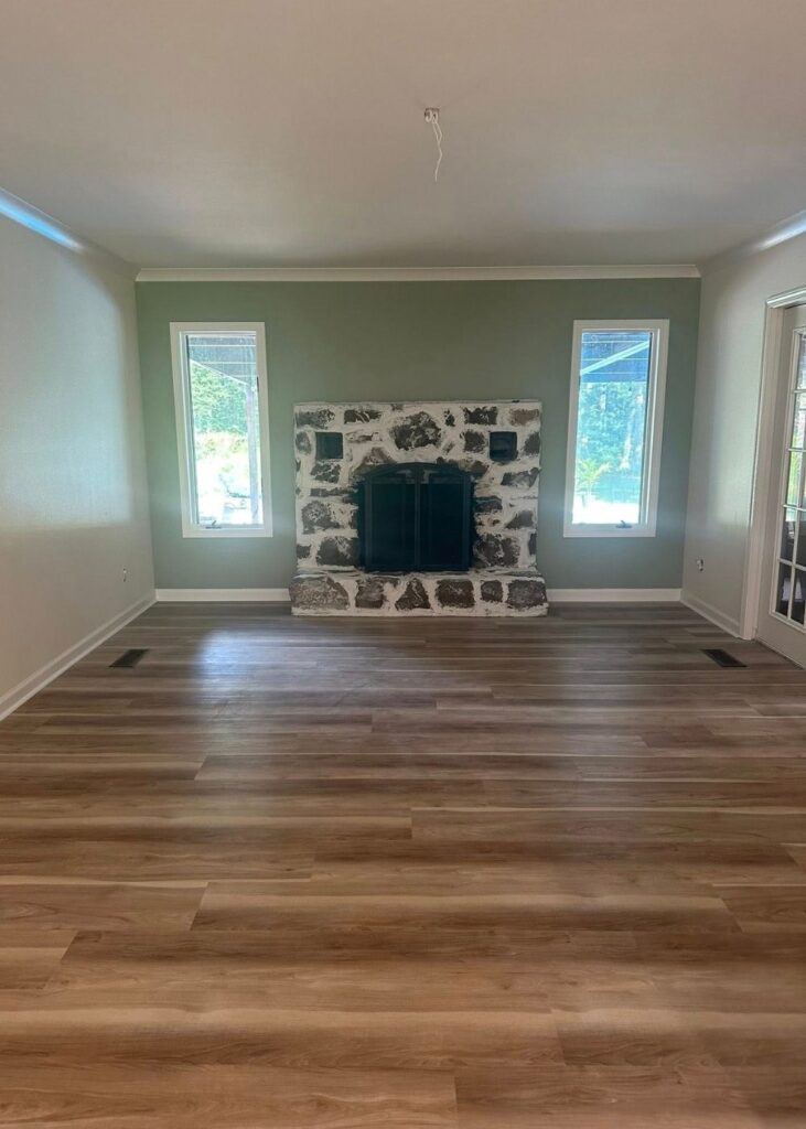 A bright, empty living room with light wood flooring, light green walls, two windows, and a stone fireplace centered between the windows. A ceiling light fixture is missing.