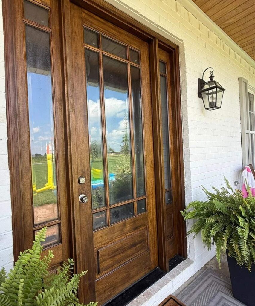 A wooden front door with glass panels set in a white brick wall, flanked by green ferns in planters. A black lantern-style light fixture is mounted beside the door, and a grassy outdoor scene is visible through the glass.