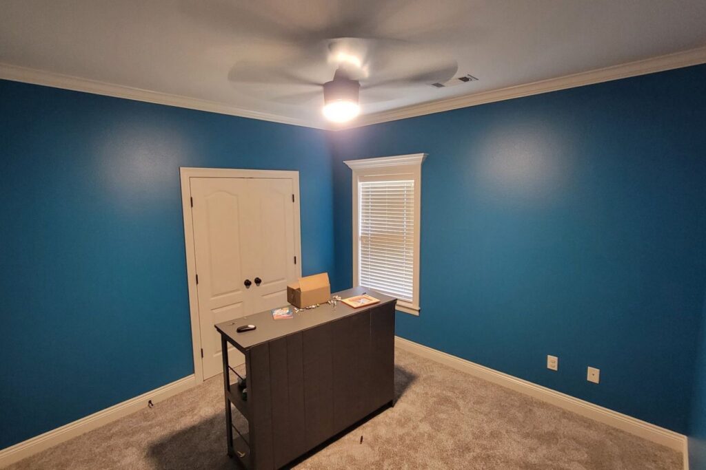 A small office room with blue walls, gray carpet, a ceiling fan light on, a window with blinds, a double-door closet, and a dark desk with office supplies.