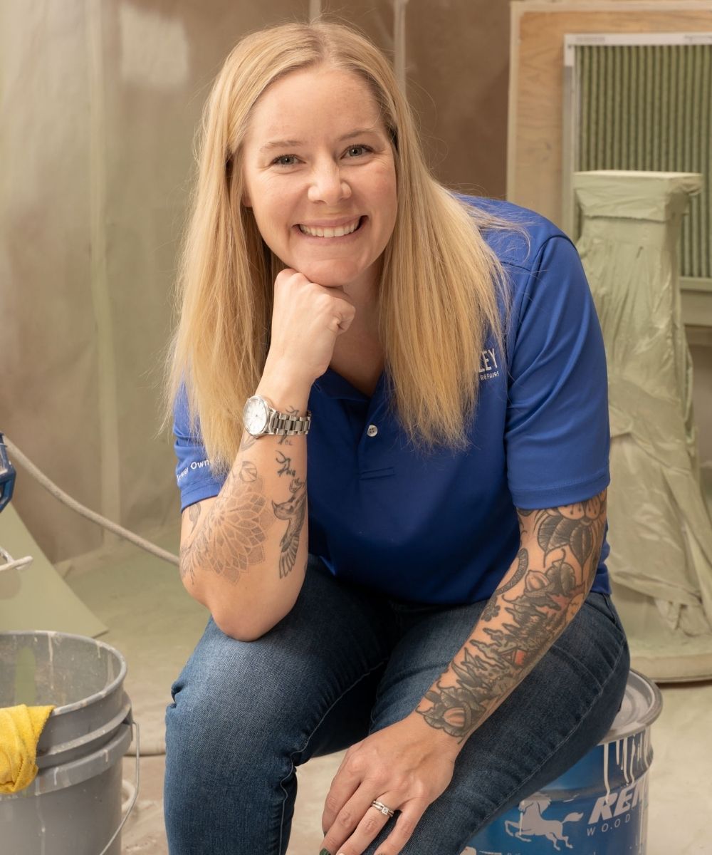 A smiling woman with long blonde hair and visible arm tattoos, wearing a blue polo shirt and jeans, leans forward with her hand on her chin in a workshop setting. Paint supplies and covered items appear in the background.