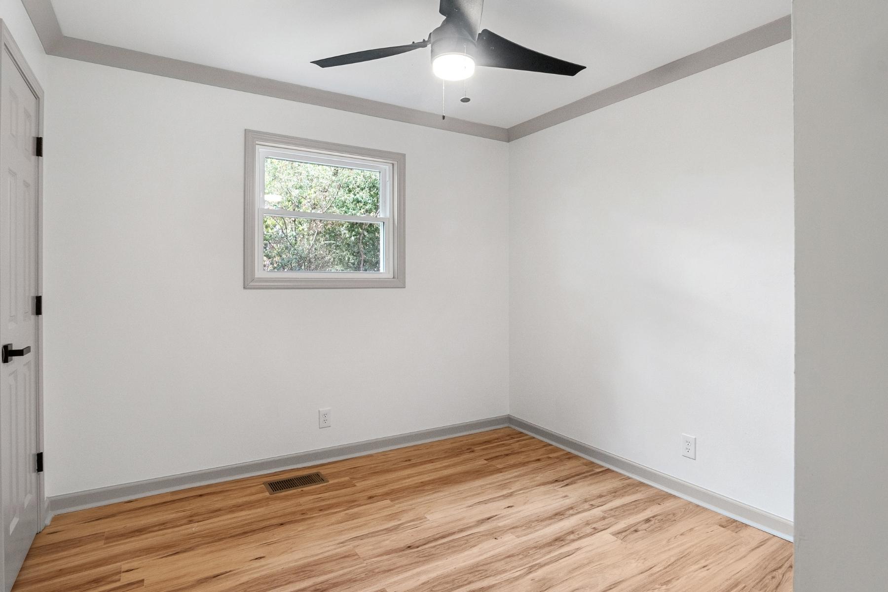 A small, empty room with light wood flooring, white walls, a single window, a black ceiling fan with a light, and gray trim around the ceiling, window, and baseboards.