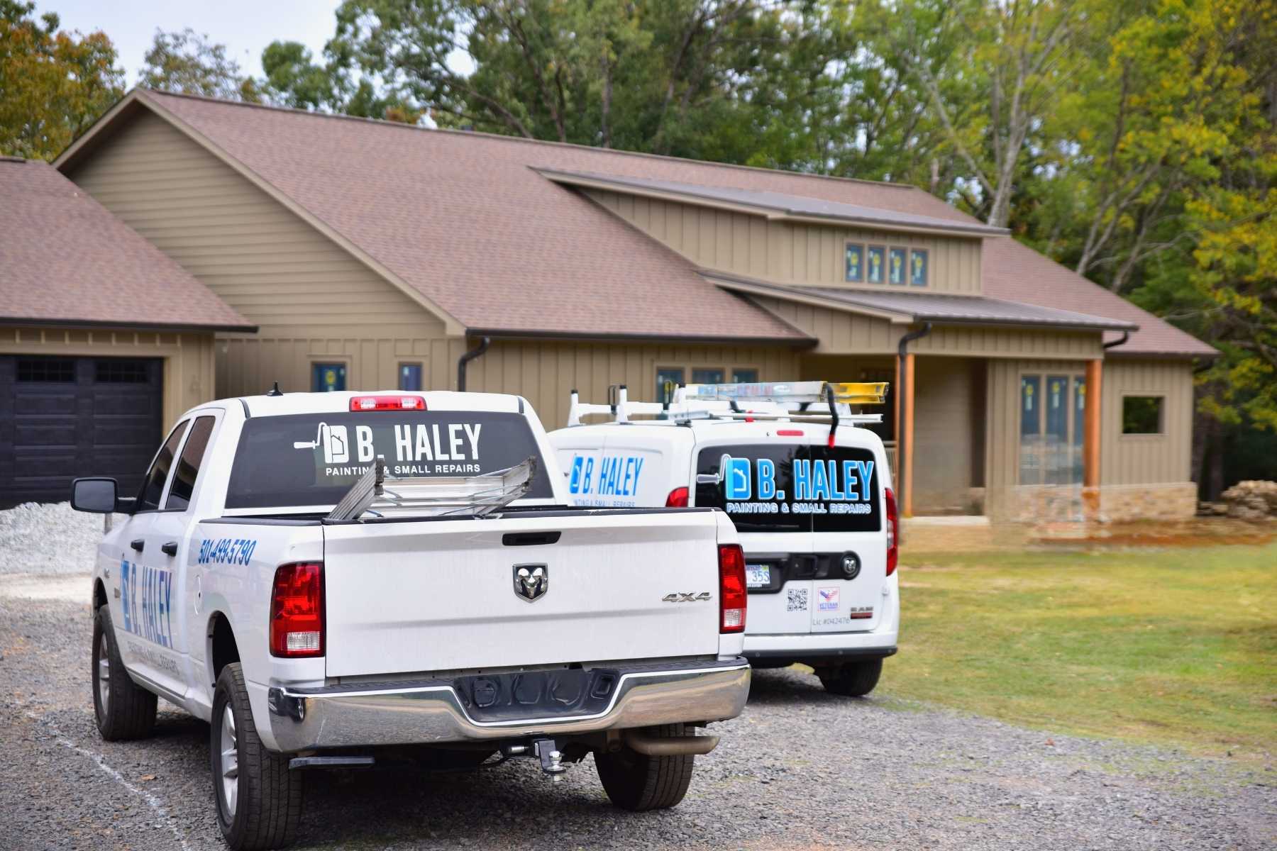 Two D.B. Haley Painting & Small Repairs vehicles, a white pickup truck and a white van, are parked in front of a beige house surrounded by trees. The vehicles display company logos and contact information.