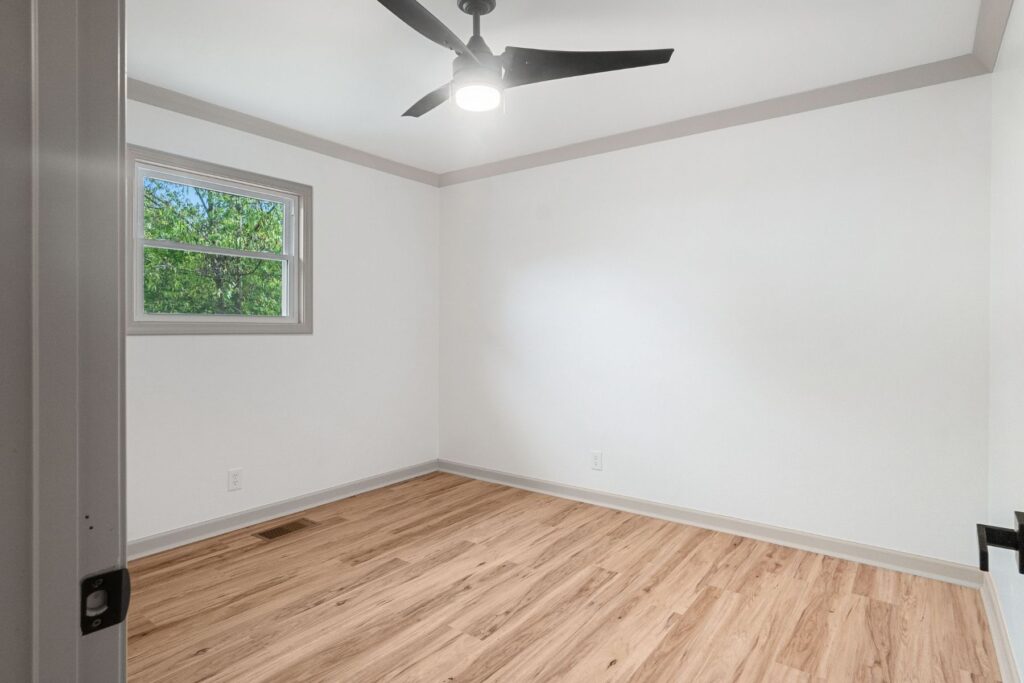 Empty room with light wood flooring, white walls, a single window showing green trees outside, a modern black ceiling fan with a light, and gray trim around the ceiling and window.