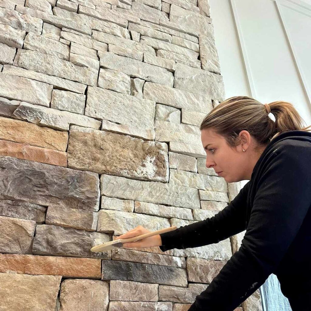 A woman in a black jacket uses a paintbrush to apply a substance to a stone wall, focusing intently on her work.