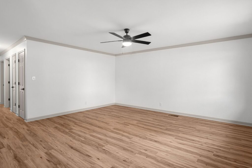 Empty room with light-colored wood flooring, white walls, and a black ceiling fan with lights. Multiple doors line the left side of the room, and the space is brightly lit with natural and overhead light.