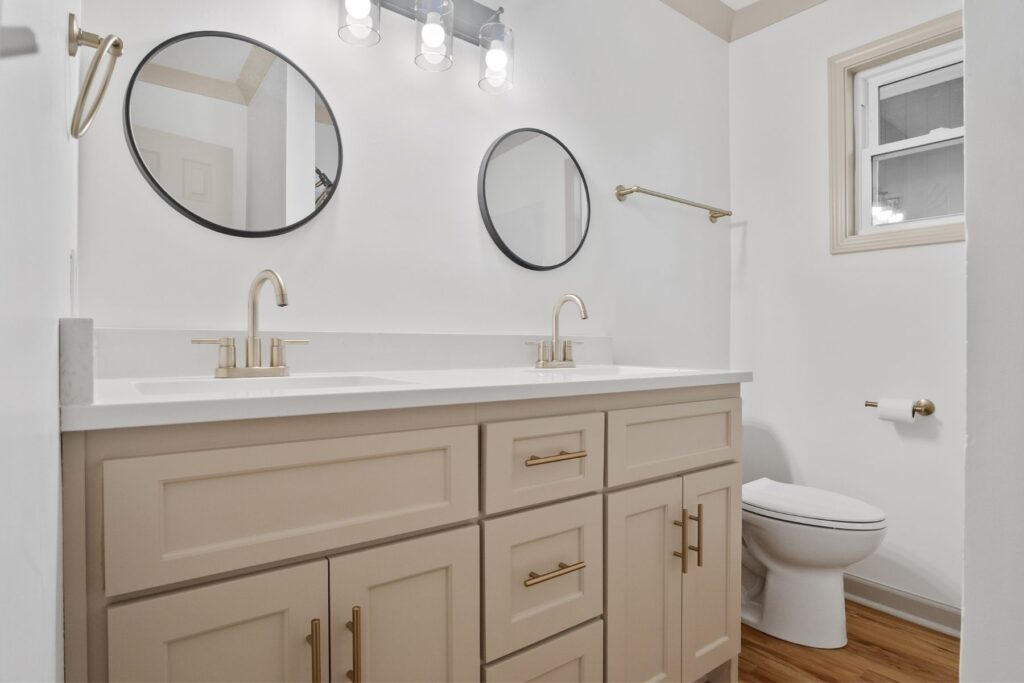 Modern bathroom with a beige double-sink vanity, gold fixtures, two round mirrors, wall-mounted lights, a toilet, wooden floor, and a small window. The walls are white with neutral trim.