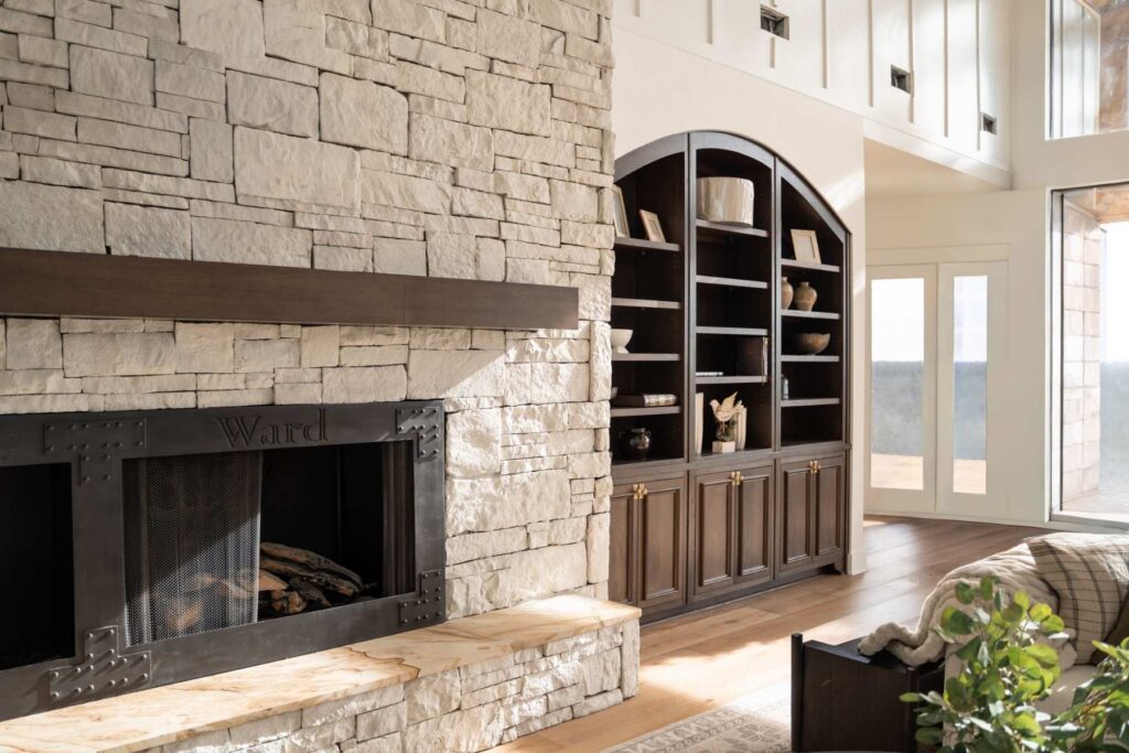 A cozy living room featuring a stone fireplace with a metal screen, built-in dark wood shelves with decor items, large windows, and natural light illuminating the space.