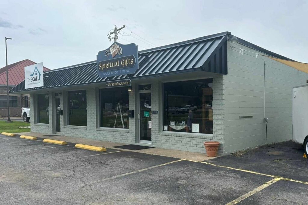 A single-story shop with large front windows and a black awning reading Spiritual Gifts above the door. The building is light gray with yellow parking lines and a sign for “The Call” on the left.
