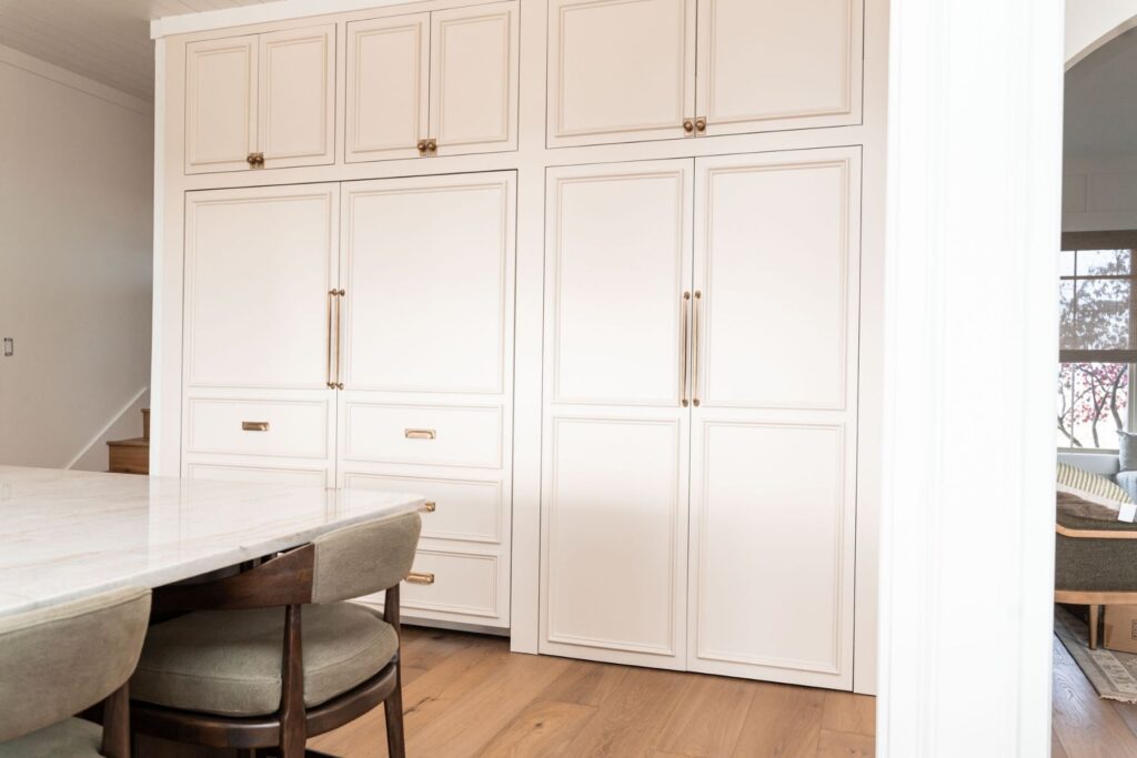 Built-in white kitchen cabinets with gold handles and drawers, next to a light-colored marble dining table with cushioned chairs on a wooden floor. Natural light enters from a window in the adjacent room.