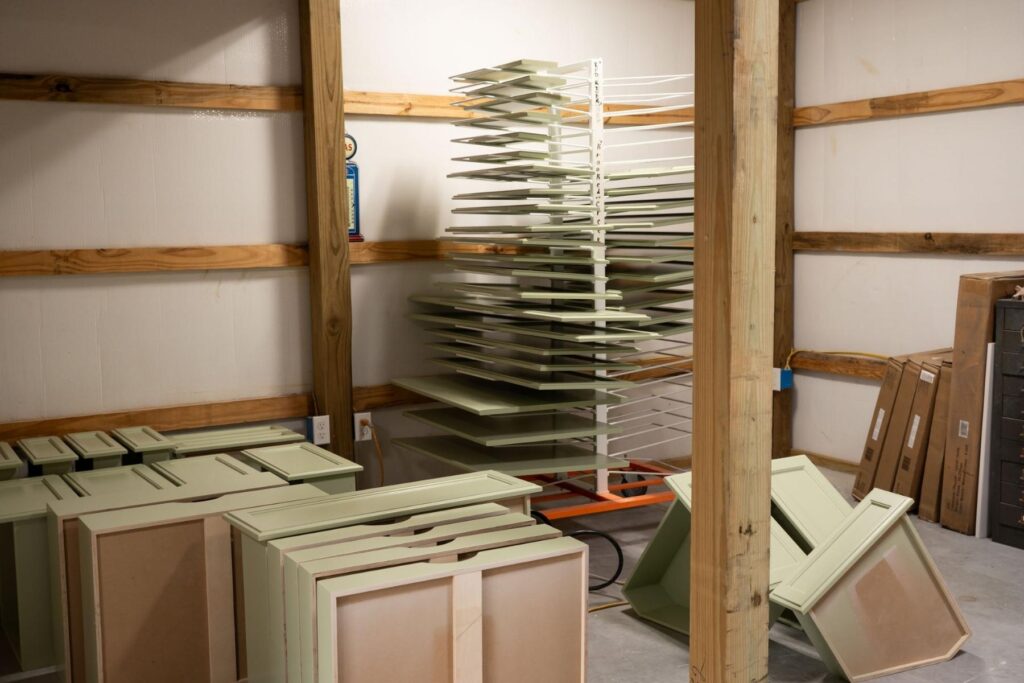 Stacked and drying painted cabinet doors and panels in a workshop, with a metal drying rack in the center, unfinished wood supports, and a fire extinguisher mounted on the back wall.