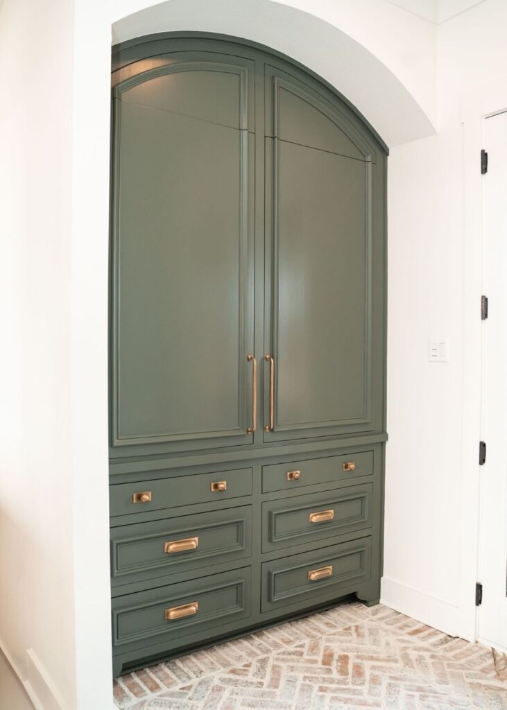 A built-in green cabinet with gold handles and multiple drawers stands under an arched alcove, beside a white wall and a white door. The floor features a herringbone brick pattern.