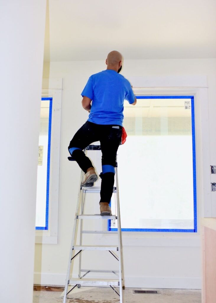 A person standing on a ladder paints the top of a wall near large windows, which are covered and taped for protection. The individual is wearing a blue shirt and black pants.