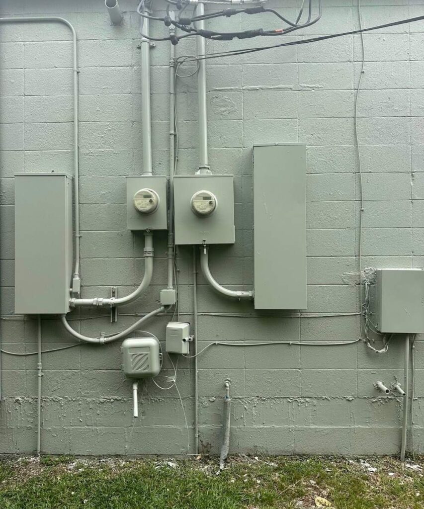Gray utility boxes, meters, and conduit pipes are mounted on a light gray concrete block wall. Wires run vertically and horizontally, with some grass visible at the bottom of the image.