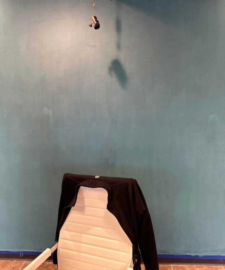 A white office chair with a black jacket draped over the back sits in front of a blue wall. Above, a small toy or object hangs from the ceiling, casting a shadow on the wall.