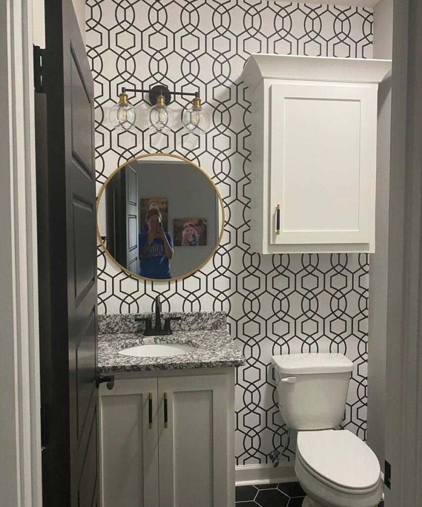 A small modern bathroom with black and white geometric wallpaper, a round mirror above a granite countertop sink, a white cabinet, a white toilet, and black hexagonal floor tiles.