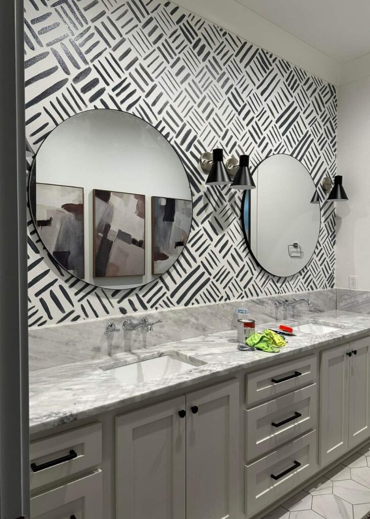 Modern bathroom with a marble countertop, two round mirrors, abstract wall art, black sconces, and geometric black-and-white wallpaper. Cleaning supplies are on the counter near one of the sinks.