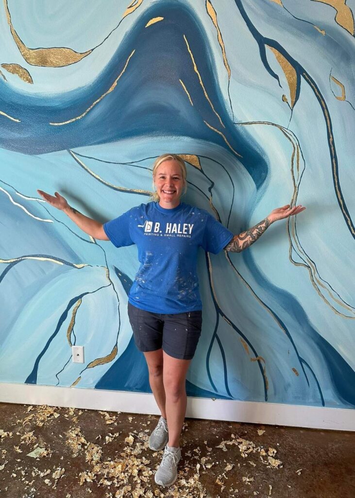 A smiling person in a blue B. Haley t-shirt, black shorts, and sneakers stands with arms outstretched in front of a blue and gold abstract mural. Paint chips are scattered on the floor.