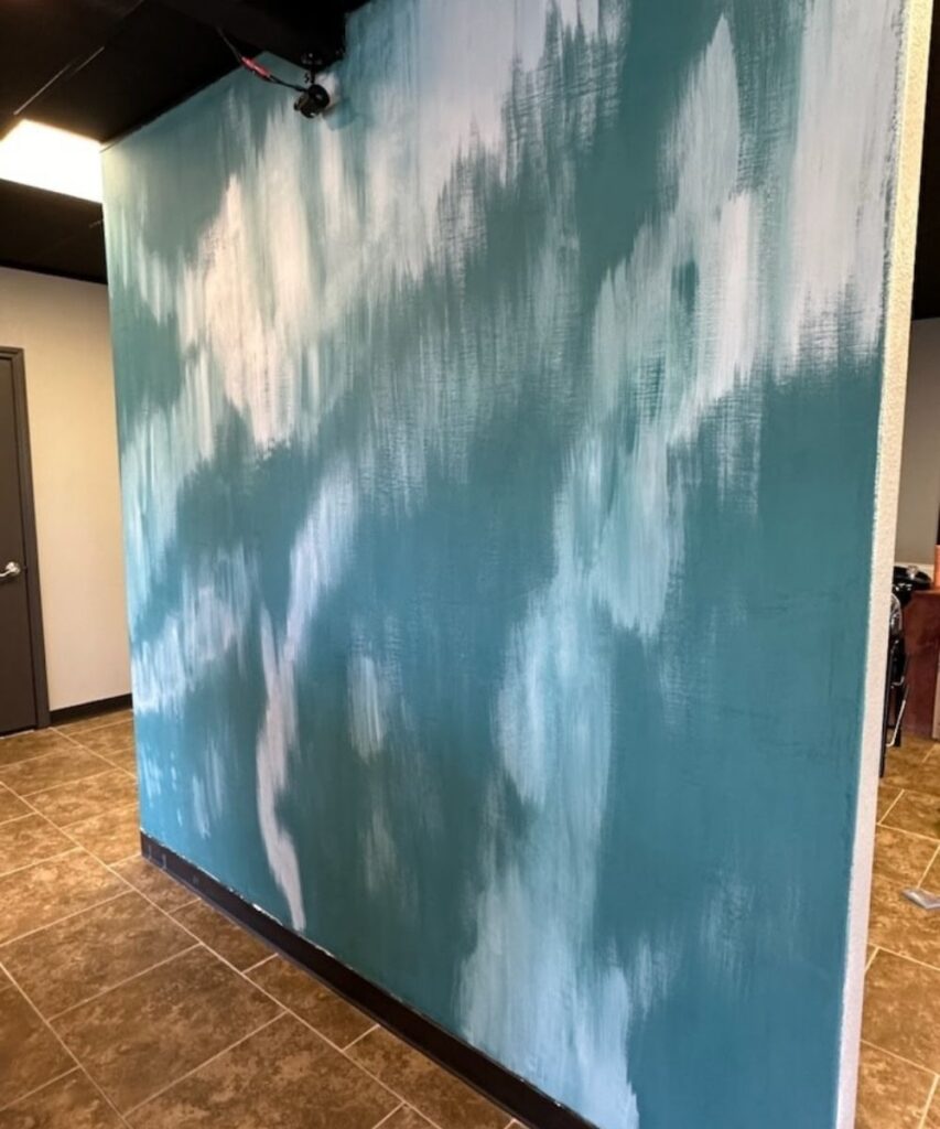 A wall painted with a teal and white abstract, brushstroke pattern stands on brown tile flooring in a room with neutral-colored walls and a black ceiling.