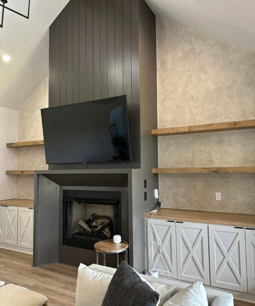 A modern living room features a large wall-mounted TV above a dark paneled fireplace, flanked by built-in shelves with wooden tops and white cabinets below, on a light wood floor.