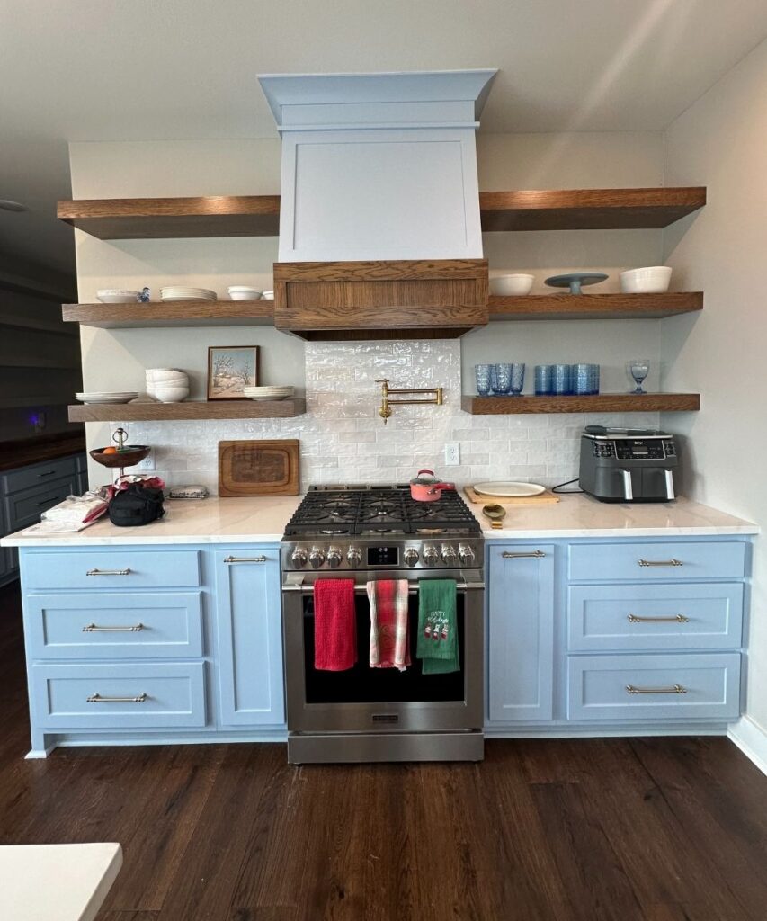 A kitchen with light blue cabinets, a stainless steel stove, wooden open shelves holding dishes, a white tile backsplash, and gold fixtures. Two towels hang from the oven, and an air fryer sits on the right counter.