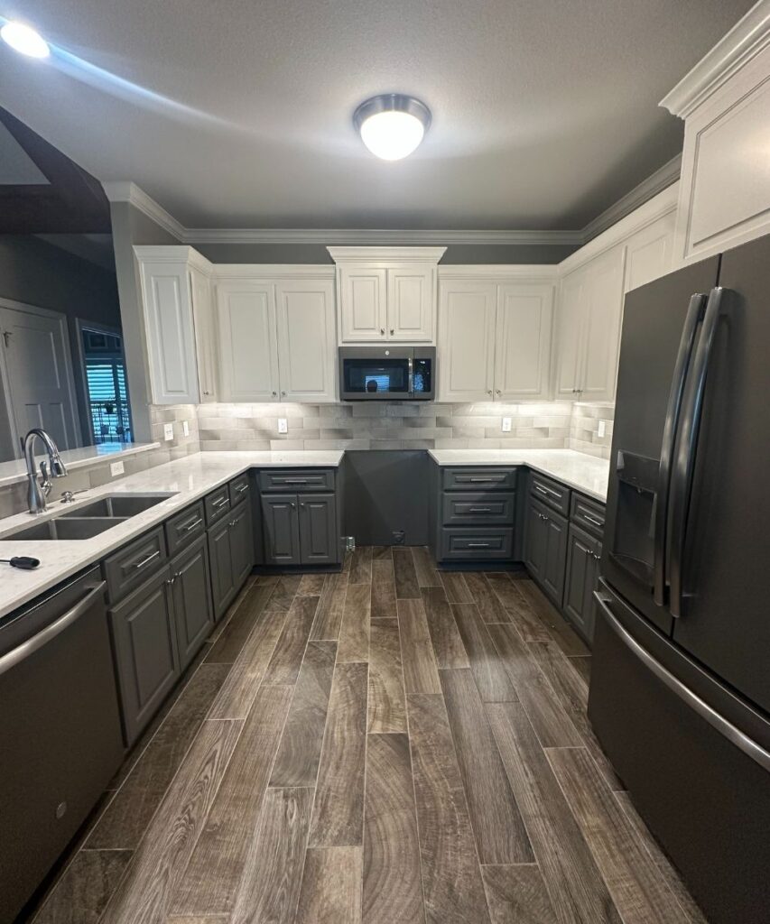 Modern kitchen with white upper cabinets, dark gray lower cabinets, stainless steel appliances, a built-in microwave, light marble countertops, gray tile backsplash, and wood-look tile flooring under bright ceiling light.