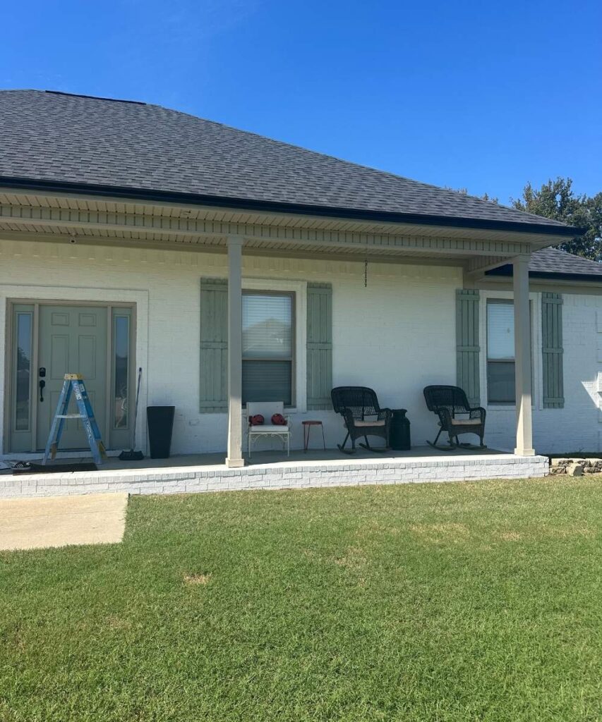 A white brick house with light green shutters and double front doors. There are chairs and a small table on the porch, a black planter, a step ladder, and a green lawn under a clear blue sky.