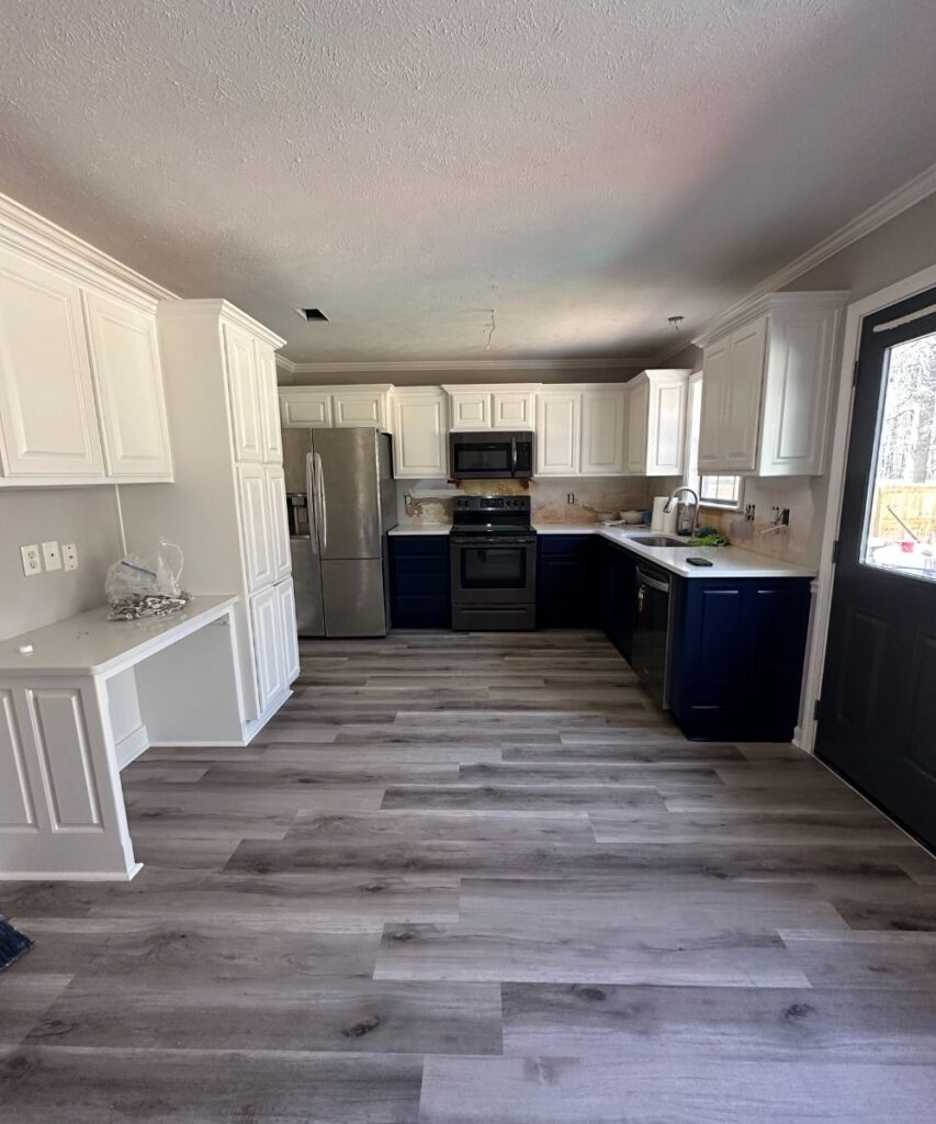Modern kitchen with white upper cabinets, dark lower cabinets, stainless steel appliances, and wood-style flooring. There’s a black door to the right and a large window letting in natural light. Counters are lightly decorated.