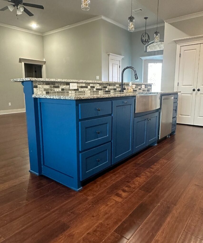 A modern kitchen features a blue island with a granite countertop, farmhouse sink, black faucet, and stainless steel dishwasher. The island contrasts with dark wood floors and light gray walls.