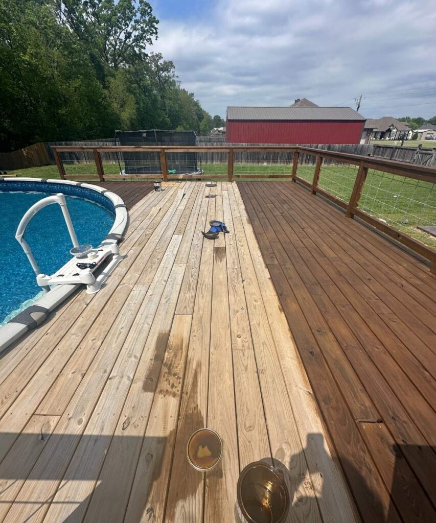 A backyard deck with a round above-ground pool on the left. The deck is partially stained, showing a contrast between treated and untreated wood. A red barn and green lawn are visible in the background under a partly cloudy sky.