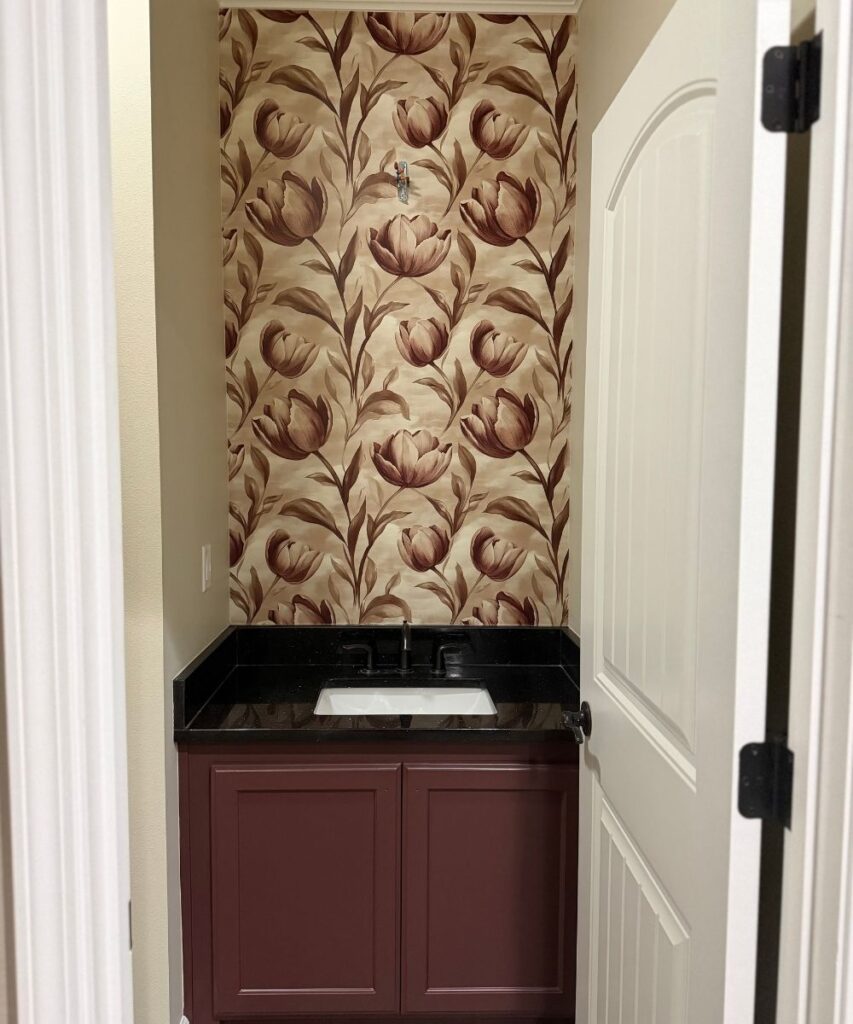 A small bathroom with burgundy cabinets, a black countertop, and a white sink. The back wall features a floral wallpaper with large, brown and beige flowers and leafy stems. The door is partially open.
