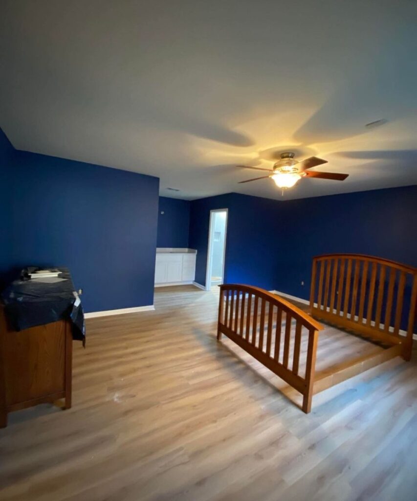 A spacious bedroom with dark blue walls, light wood flooring, a wooden bed frame, ceiling fan with lights, a dresser on the left, and a white vanity with a mirror in the back corner near a door.