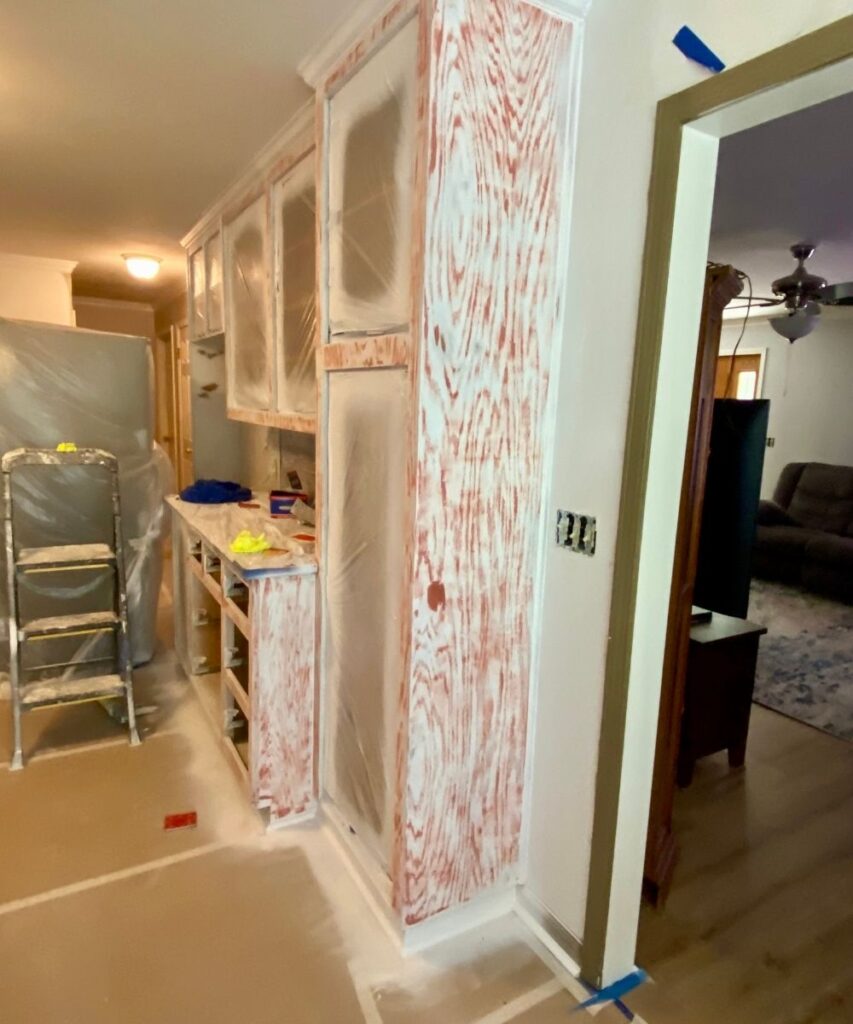 Kitchen cabinets covered in plastic and red-tinted primer during a renovation; floor and surrounding areas are protected with paper and tape. A ladder and tools are nearby, and the room looks mid-renovation.