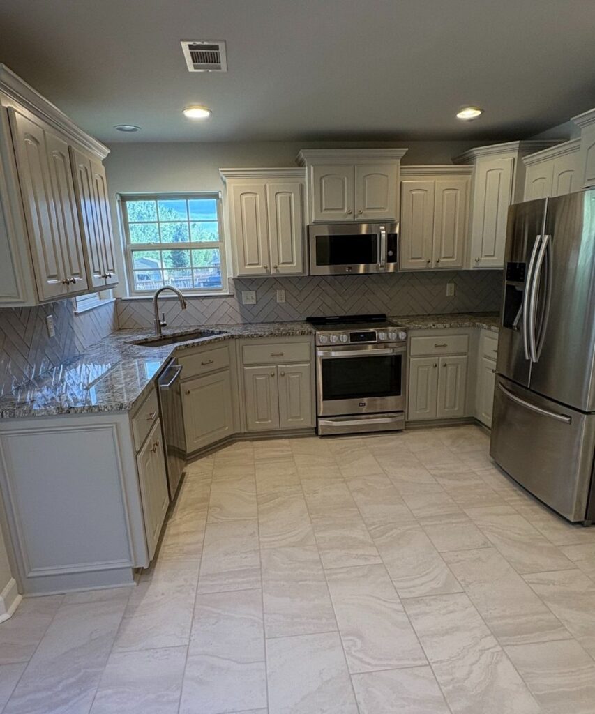 Modern kitchen with white cabinets, stainless steel appliances, a gas stove with oven, microwave, double-door fridge, granite countertops, tile backsplash in a herringbone pattern, and a window above the sink.
