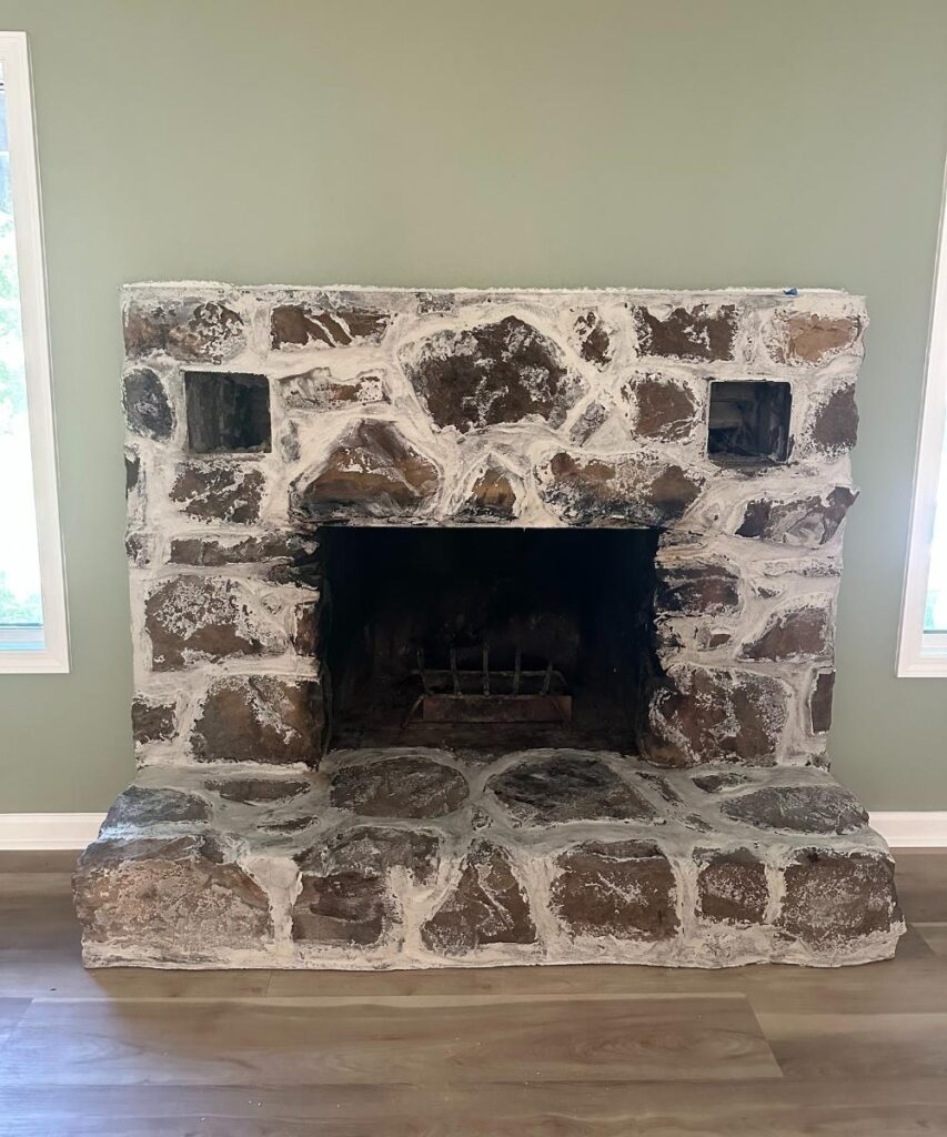 A rustic stone fireplace with a dark firebox, two small openings above, and a wide stone hearth, set against a light green wall with wood flooring.