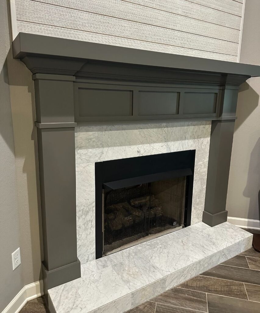 A modern fireplace with a white marble surround, gray mantel, and decorative molding, set against a wall with light wood paneling above, and dark wood floor tiles below.