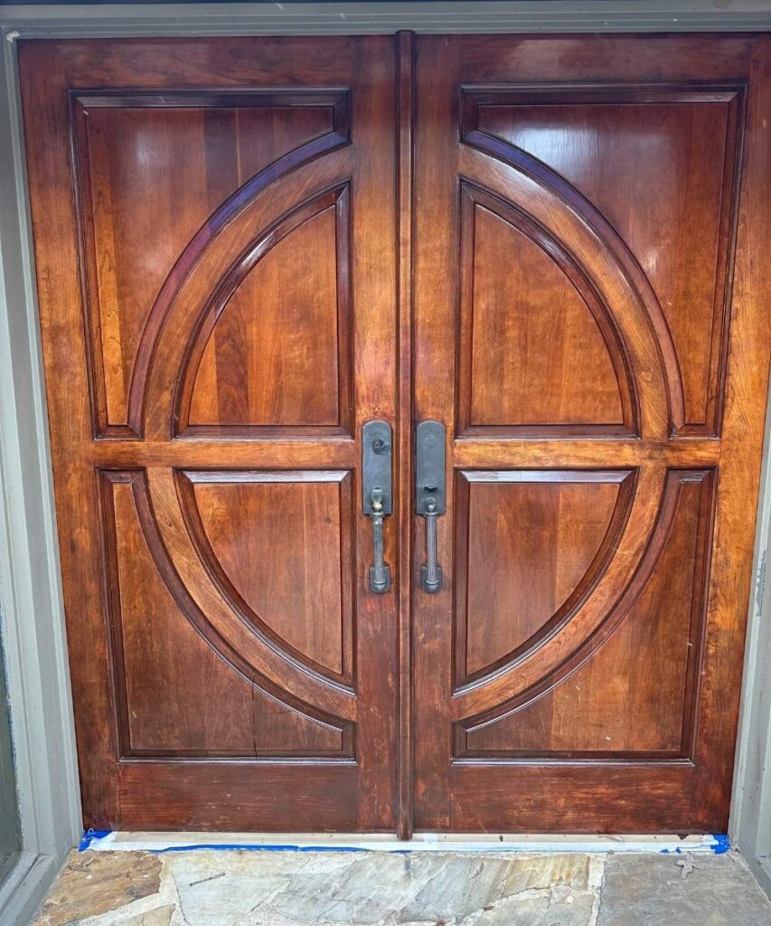 A set of double wooden doors with a rich, dark stain and decorative circular carvings. Each door has a vertical handle and lock, and the doors are framed by stone flooring at the bottom.
