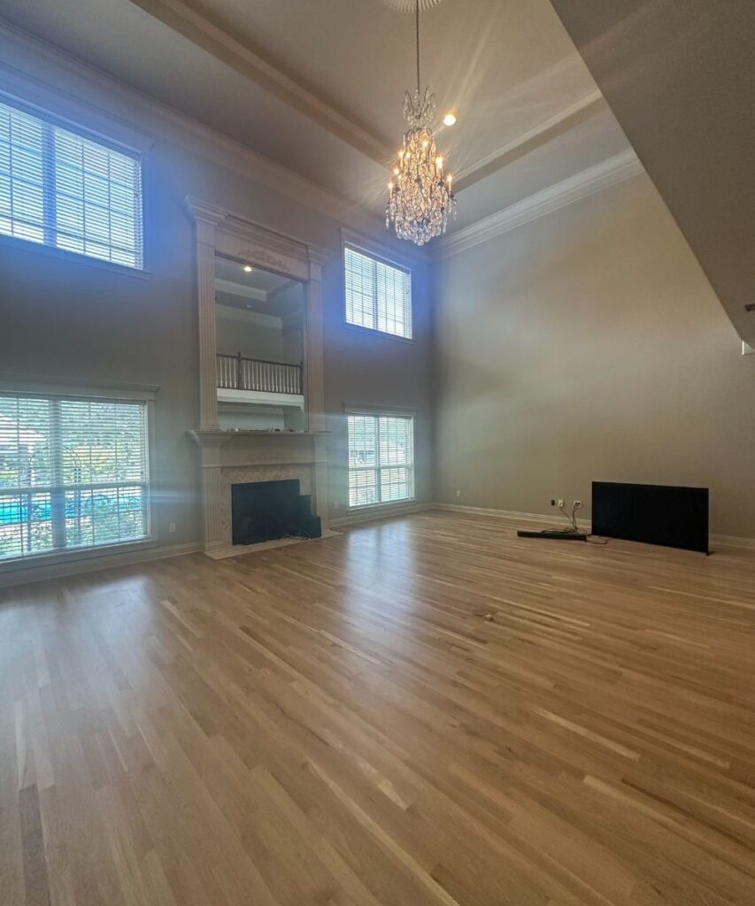 Spacious, empty living room with light wood floors, tall ceilings, large windows, a chandelier, a fireplace with a mirror above, and a TV placed on the floor against the wall. Natural light fills the room.