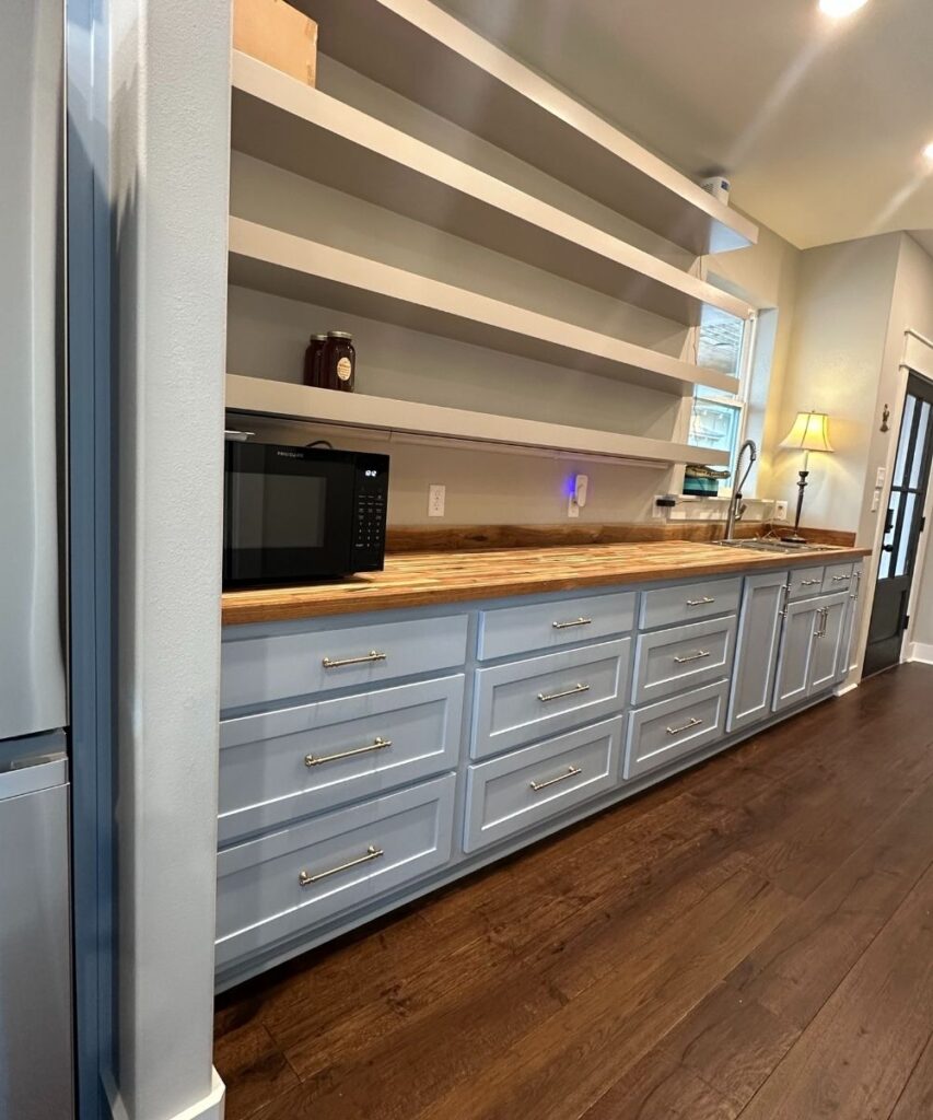 A kitchen with wooden floors, light blue lower cabinets with silver handles, open wooden shelves, a microwave on the countertop, a lamp, and various items on the shelves.