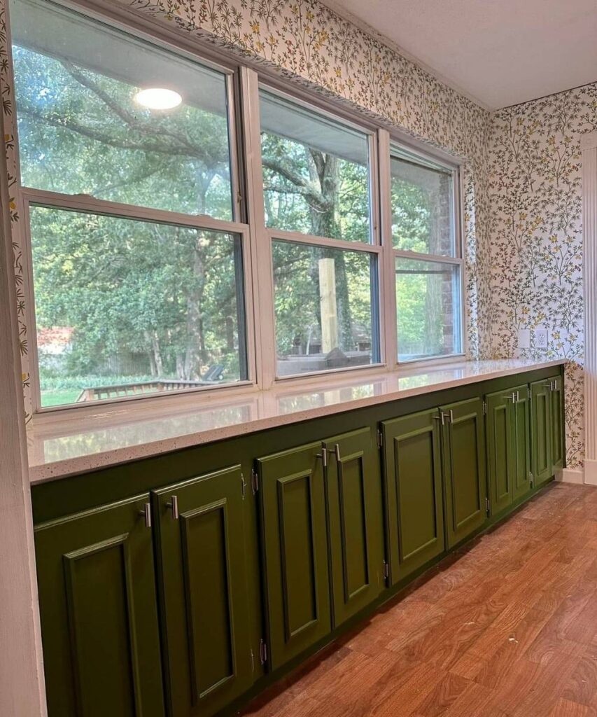 A row of green cabinets with silver handles is set against a wall with large windows and nature-inspired wallpaper. Sunlight streams in, illuminating the wood floor and the outdoor greenery seen through the windows.