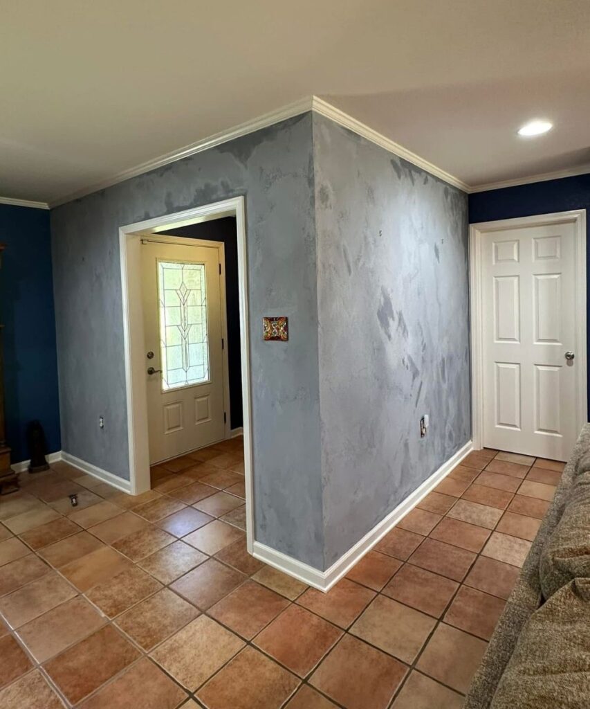 A hallway with beige and brown tiled floor, light gray textured walls, white trim, a white door at the end, and a front door with a glass window on the left. A couch is partially visible on the right.