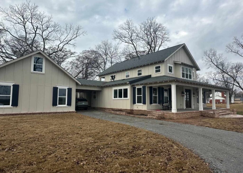 A two-story beige house with dark shutters and a covered porch sits on a brown lawn with bare trees in the background under a cloudy sky. A driveway curves to the left, leading to a carport attached to the house.