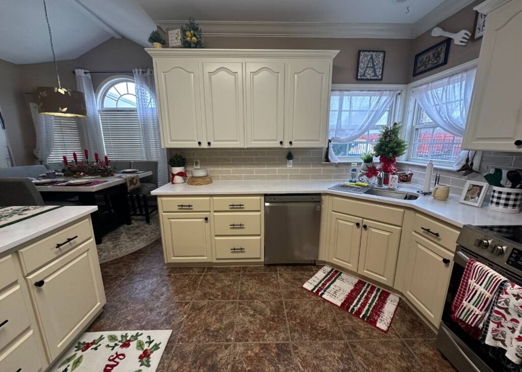 A cozy kitchen with cream cabinets, stainless steel appliances, Christmas-themed rugs, and holiday decorations. Windows with sheer curtains let in natural light, and a dining area is visible on the left.